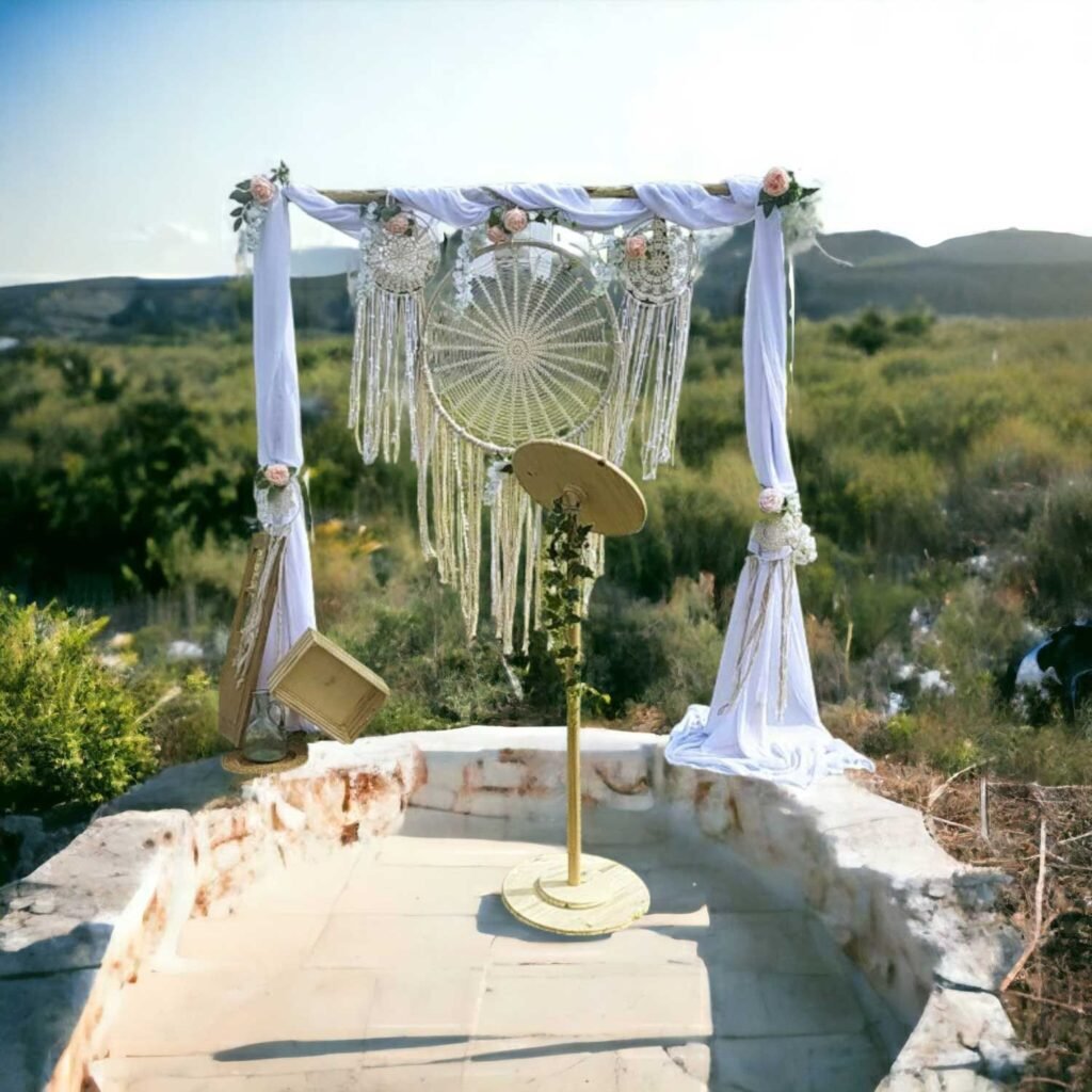 A wooden wedding arch decorated with white veils and white flower garlands with green foliage, set outdoors near a pond. In front of the arch, two folding wooden chairs are placed next to a wooden lectern, also dressed with greenery. The whole scene is surrounded by natural vegetation, with trees in the background under a clear sky.