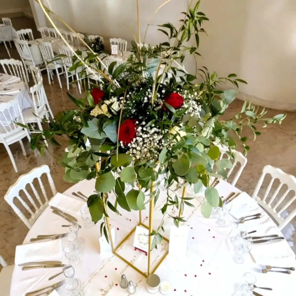 Elegant floral centrepiece for a Var wedding, composed of red and white roses, baby's breath and green foliage, set on a gold stand. The table decor is completed with white chairs and a white tablecloth, creating a refined, romantic mood for the reception. Ideal for a red-and-white themed wedding with fresh flowers.