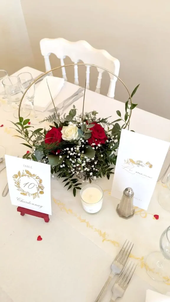 Floral wedding centerpiece with red and white roses, accompanied by gypsophila and foliage, displayed in a gold hoop on an elegant table. The decoration also includes a menu card and a table number card marked 'Table 10 Chardonnay', all on a white tablecloth with a refined, romantic atmosphere. Ideal for a wedding in the Var with a classic, floral theme.