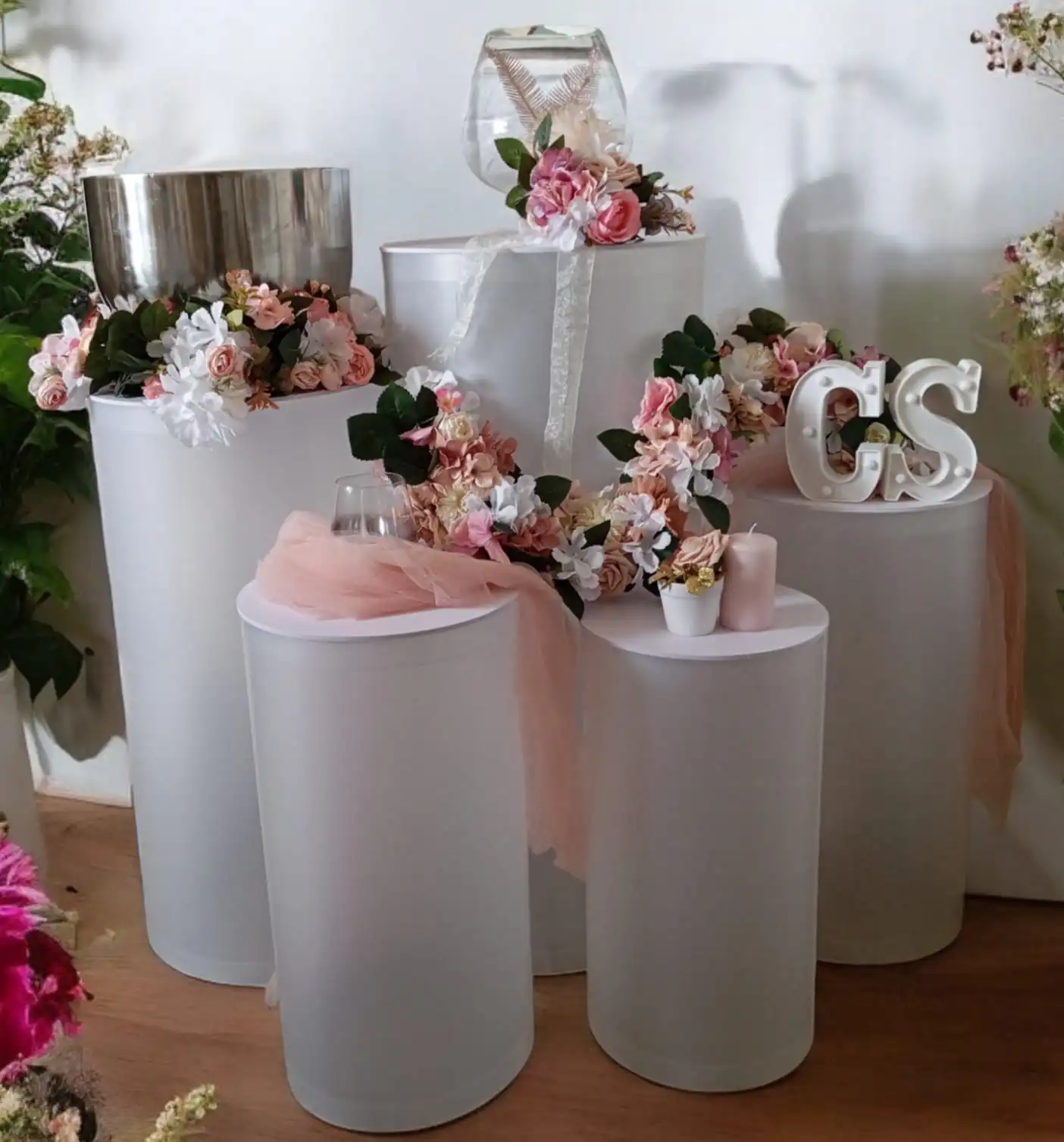 Wedding cylindrical column: wedding decor arrangement with white and clear columns. The columns are decorated with artificial flowers in pink, white and green tones. Pink candles and a pink tulle scarf add a romantic touch. A large glass vase and decorative “CS” letters also feature on the columns.