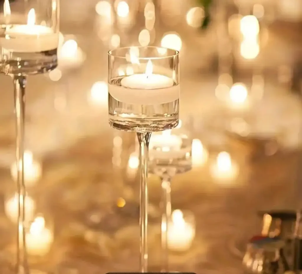 Photophore en verre transparent avec bougie flottante allumée, placé sur une table de réception. Le photophore est sur pied, apportant une ambiance chaleureuse et élégante grâce aux multiples reflets de lumière sur la table et les autres bougies en arrière-plan. L'atmosphère est tamisée et romantique, idéale pour une décoration de mariage.