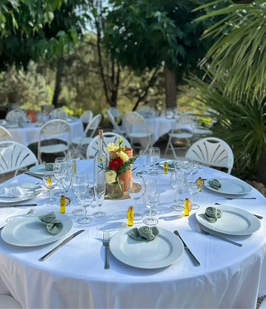 L'image montre une table de mariage élégamment dressée en extérieur, sous des arbres verdoyants. La table ronde est recouverte d'une nappe blanche immaculée et est dressée avec soin pour chaque invité. Au centre, un bouquet de fleurs coloré, composé de roses blanches et de fleurs rouges, est placé dans un vase, accompagné d'une bouteille en verre transparente, ce qui apporte une touche de fraîcheur et de naturel à la décoration. Chaque assiette est ornée d'une serviette roulée de couleur vert pastel, et de petites bouteilles d'huile d'olive sont placées près des assiettes, ajoutant un charme méditerranéen à l'ensemble. Les chaises blanches autour de la table complètent cet environnement harmonieux et accueillant, parfait pour une réception de mariage en plein air.