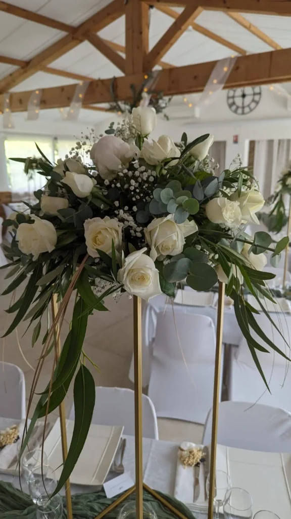 Tall floral centerpiece on a gold-tone structure with a bouquet of white roses, eucalyptus and gypsophila. Natural, luxurious wedding decoration, cascading foliage, romantic and verdant atmosphere under a wooden frame, tables draped in white and refined decoration.