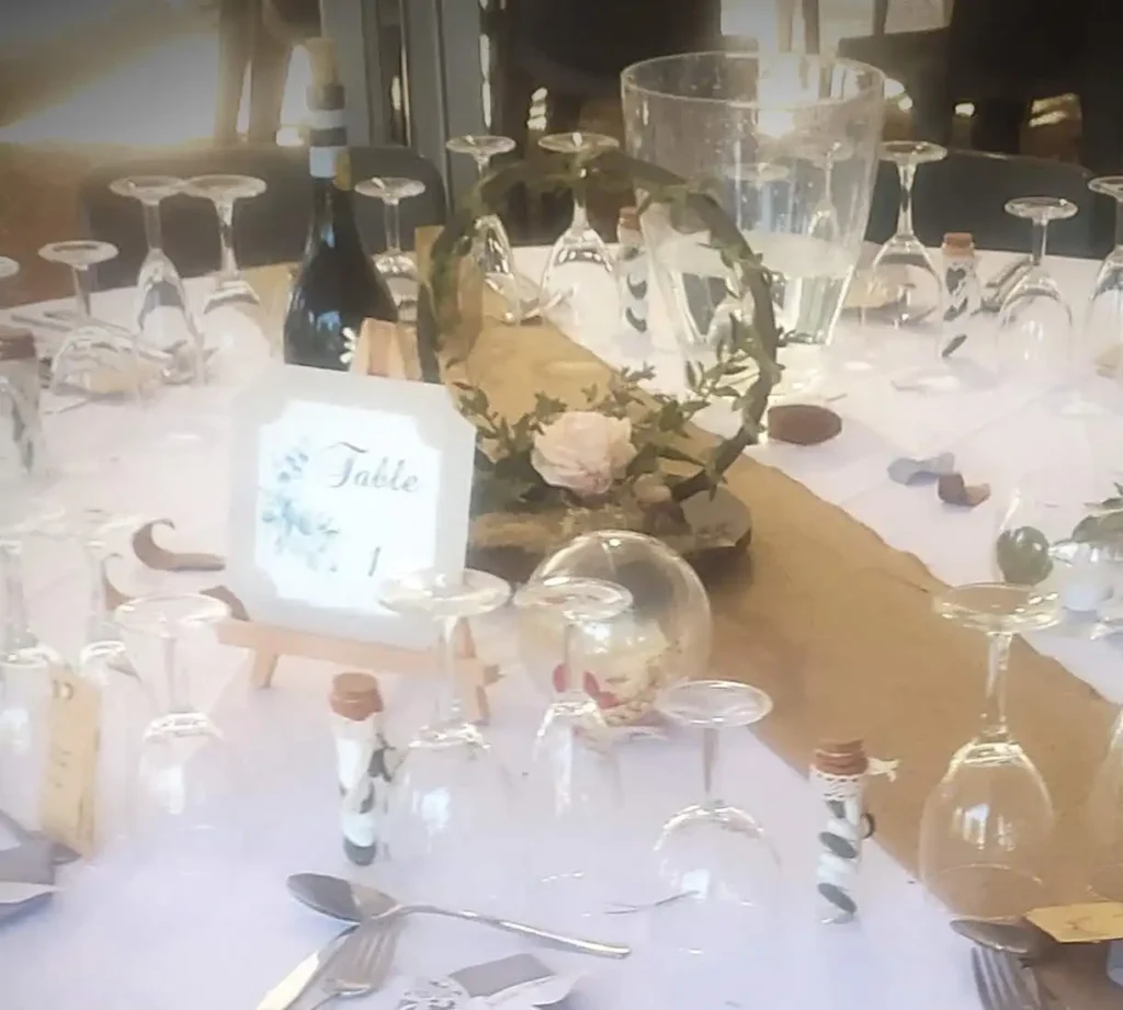 Rustic, countryside wedding table decor with jute table runner, clear tableware, upturned glasses and wood logs. Centrepiece composed of a botanical wreath dressed with foliage, ivy and powder-pink roses — a soft, authentic atmosphere for an elegant countryside reception.