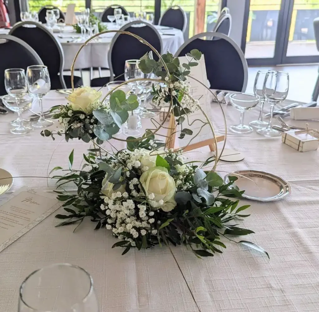 Round wedding centerpiece, modern floral arrangement with gold hoops, white roses, eucalyptus and gypsophila. Elegant arrangement on an ivory tablecloth, romantic and contemporary atmosphere, perfect for a natural chic or bohemian wedding.