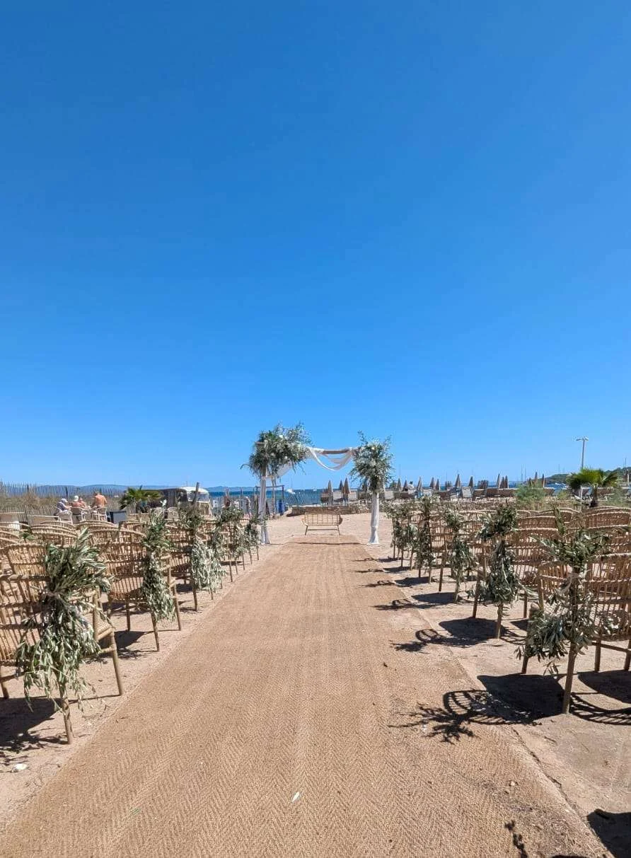 Allée de cérémonie laïque sur la plage, bordée de chaises en rotin décorées de feuillage d’olivier et d’une arche habillée de voilage blanc face à la mer.