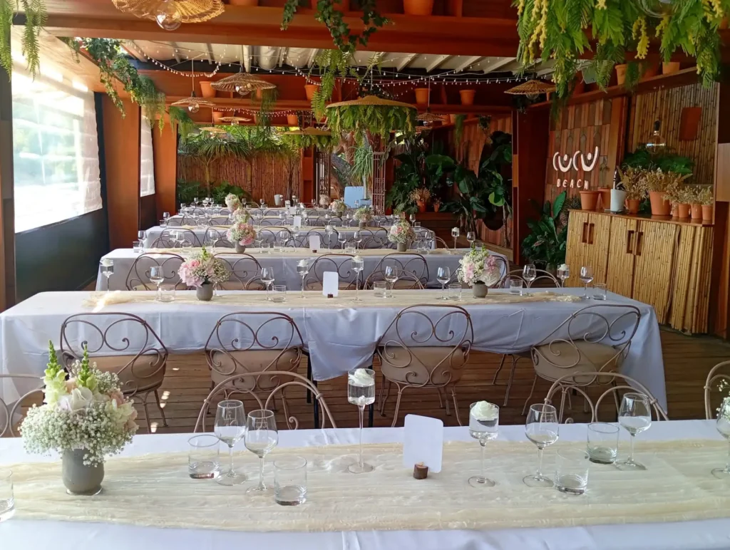 Boho, natural wedding reception room decorated with rectangular tables, floral centrepieces, stoneware vases, baby's breath, pastel roses and foliage. Tropical, warm atmosphere with rattan, wood, bamboo suspensions and hanging plants — a chic, nature-inspired pop-up bar spirit.