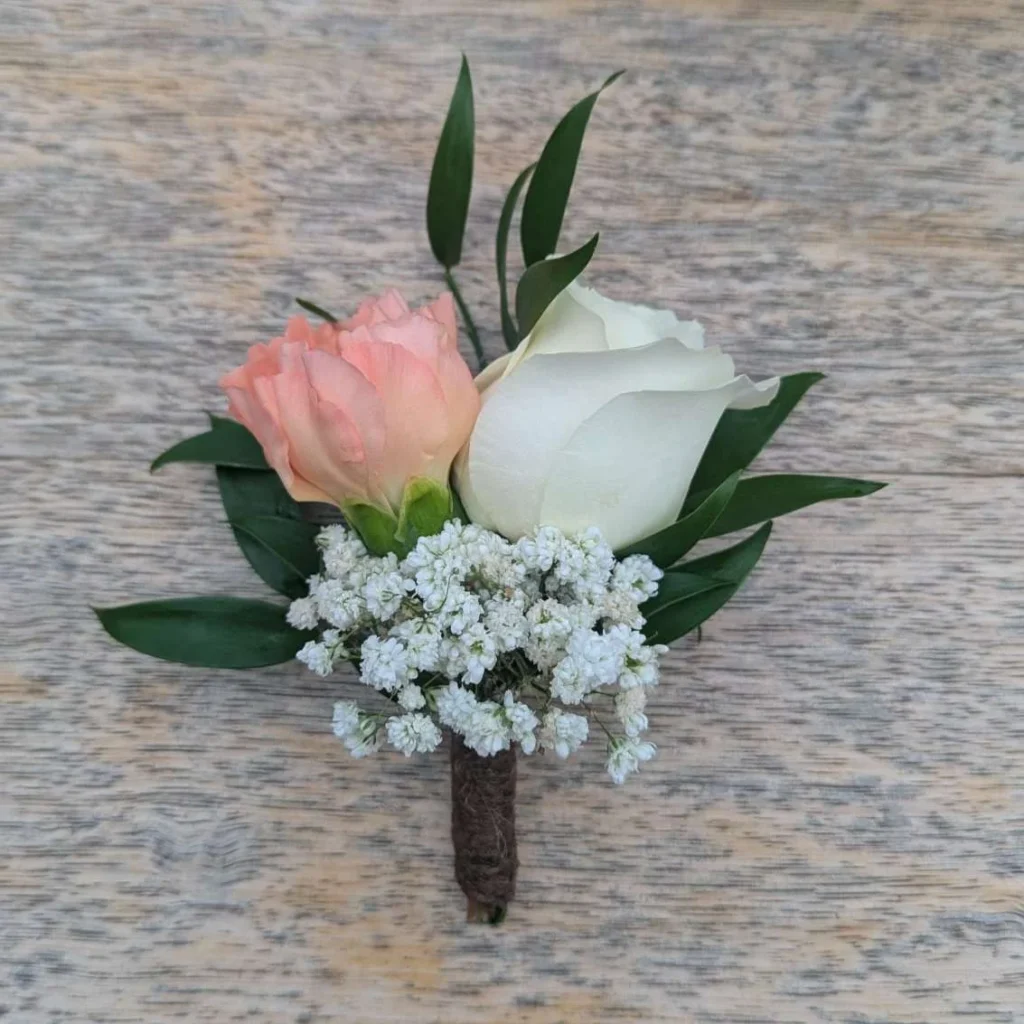 Wedding boutonnière composed of a white rose, a peach rose, white gypsophila and green foliage, mounted on a stem and set on a wooden table.