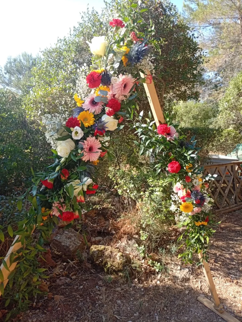 Triangular wooden wedding arch adorned with colourful fresh flowers, gerberas, carnations and blue thistles - Iliade Création Var